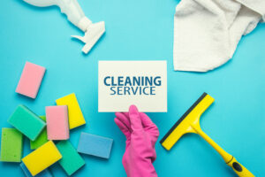 Female hand with pink gloves, holding a card with text Cleaning Services and accessories for cleaning on a blue background. cleaning service concept. Flat lay, Top view.